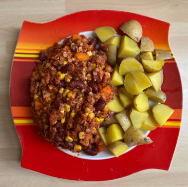 Vegetarisches Chili mit Kidneybohnen, Mais, Gemüse und Kartoffeln auf einem roten Teller.